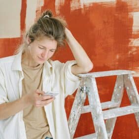 En person lener seg mot en skrittstige foran en vegg med rødbrun maling, mens de ser på smarttelefonen sin.