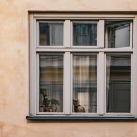 Et vindu i en lys ferskenfarget vegg med en brunstripet katt som sitter i vinduskarmen.